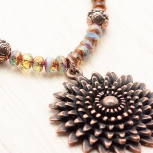 Puede incluir: Un collar de tono cobre con un gran colgante de girasol adornado. El collar est&aacute; hecho con cuentas de vidrio iridiscente y cuentas de tono cobre.