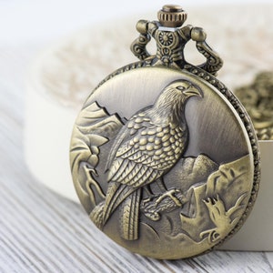 May include: A vintage-style pocket watch with a bronze finish. The watch face features an engraved image of a bird with detailed feathers and a mountain scene.