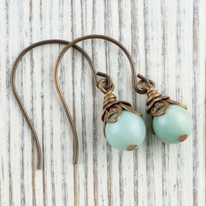 May include: A pair of earrings with a bronze-colored hook and decorative top. Each earring features a light blue, round bead. The earrings are displayed on a white, weathered wooden surface.