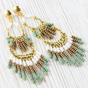 May include: A pair of long, gold-toned earrings with white, green, and brown beads. The earrings have a hammered metal design and dangle from the earlobe.