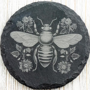 Op de afbeelding: Een zwarte leisteen onderzetter met een lasergegraveerd ontwerp van een bij omringd door bloemen. De bij is grijs en heeft een gestreepte buik. De bloemen zijn wit en grijs.