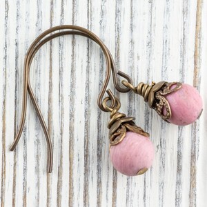 K&ouml;nnte beinhalten: Handgefertigte Ohrringe mit rosa, ovalen Perlen und antiken bronzefarbenen Metallarbeiten. Die Ohrhaken sind ebenfalls bronzefarben. Die Ohrringe werden auf einer wei&szlig;en, verwitterten Holzoberfl&auml;che pr&auml;sentiert.
