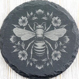 May include: Round, dark gray slate coaster with an engraved design of a bee surrounded by flowers and leaves. The bee is centered, with detailed wings and body. The floral elements create a decorative border around the bee.