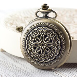 K&ouml;nnte beinhalten: Antike bronzefarbene Taschenuhr mit einem verzierten Blumendesign auf der Vorderseite. Die Uhr hat eine runde Form mit einem detaillierten, durchbrochenen Muster. Sie befindet sich vor einem wei&szlig;en Hintergrund.