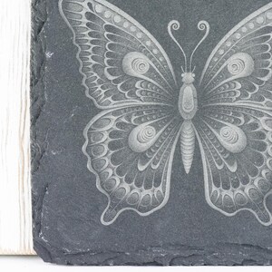 Puede incluir: Un posavasos de pizarra negra con un dise&ntilde;o de mariposa grabado en blanco. La mariposa tiene detalles intrincados y una apariencia delicada.