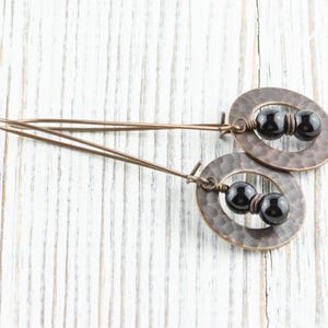 Peut inclure: Boucles d'oreilles artisanales avec des pendentifs ovales en cuivre martel&eacute;. Chaque pendentif contient deux perles noires. Les boucles d'oreilles ont un design simple et &eacute;l&eacute;gant, parfait pour un usage quotidien. Elles sont pr&eacute;sent&eacute;es sur une surface en bois blanc.
