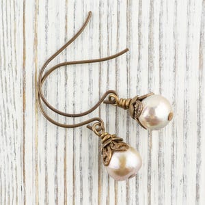 May include: A pair of drop earrings with pearlescent, teardrop-shaped pearls. The earrings have bronze-colored, decorative caps and ear wires. The earrings are displayed on a white, weathered wooden surface.