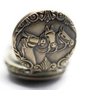 Puede incluir: Un reloj de bolsillo dorado con una cara circular que presenta un dise&ntilde;o en relieve de tres cabezas de caballo. Los caballos est&aacute;n mirando en la misma direcci&oacute;n y sus cabezas se tocan.