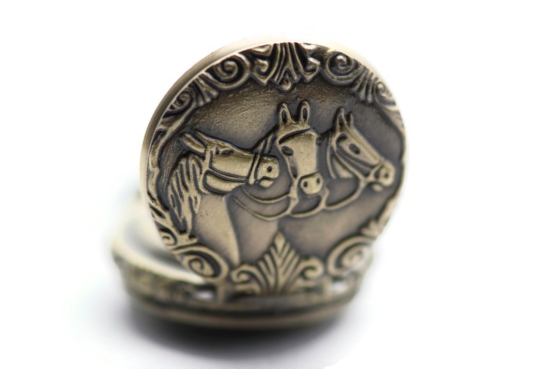 Puede incluir: Un reloj de bolsillo dorado con una cara circular que presenta un dise&ntilde;o en relieve de tres cabezas de caballo. Los caballos est&aacute;n mirando en la misma direcci&oacute;n y sus cabezas se tocan.