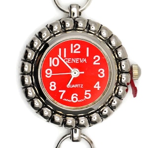 Puede incluir: Un reloj de bolsillo plateado con una esfera roja y agujas negras. El reloj tiene un bisel con cuentas y un lazo en la parte superior para sujetarlo a una cadena. La esfera del reloj dice "GENEVA QUARTZ" y tiene números negros.