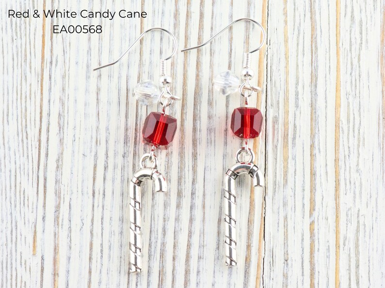 Pode incluir: Um par de brincos com tema natalino. Cada brinco apresenta uma conta vermelha facetada, um pingente de bengala doce prateado e um detalhe de cristal transparente. O texto "Red & White Candy Cane EA00568" &eacute; vis&iacute;vel.