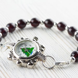 May include: A silver watch with a white face and a Christmas tree design. The watch is attached to a bracelet made of dark red beads.