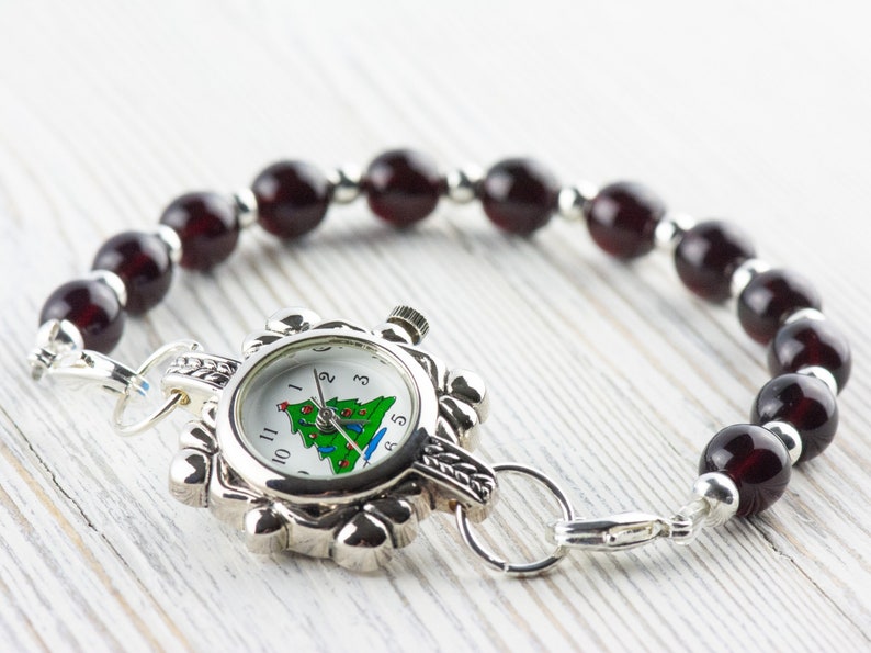 May include: A silver watch with a white face and a Christmas tree design. The watch is attached to a bracelet made of dark red beads.