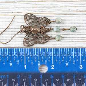 May include: A bronze-colored butterfly-shaped earring with intricate filigree details. The earring features three dangling beads in shades of green and white. The earring measures approximately 1.5 inches in length.