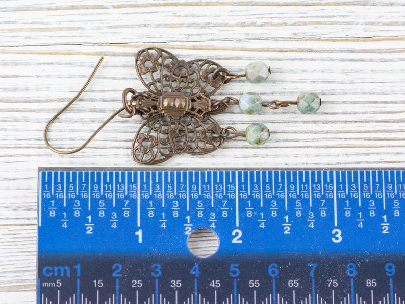 May include: A bronze-colored butterfly-shaped earring with intricate filigree details. The earring features three dangling beads in shades of green and white. The earring measures approximately 1.5 inches in length.
