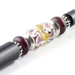 K&ouml;nnte beinhalten: Ein schwarzer Stift mit einem dekorativen Design aus lila, wei&szlig;en und goldenen Perlen. Die Perlen sind in einem Muster angeordnet und mit silbernem Metall am Stift befestigt.