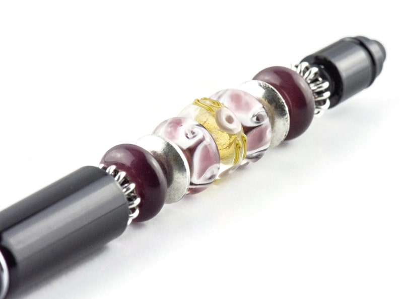 K&ouml;nnte beinhalten: Ein schwarzer Stift mit einem dekorativen Design aus lila, wei&szlig;en und goldenen Perlen. Die Perlen sind in einem Muster angeordnet und mit silbernem Metall am Stift befestigt.