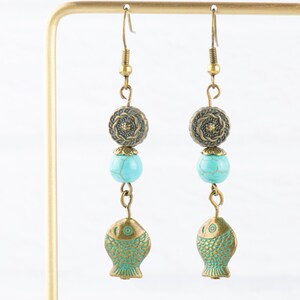 May include: A pair of turquoise and brass dangle earrings featuring a turquoise bead, a brass flower charm, and a brass fish charm.