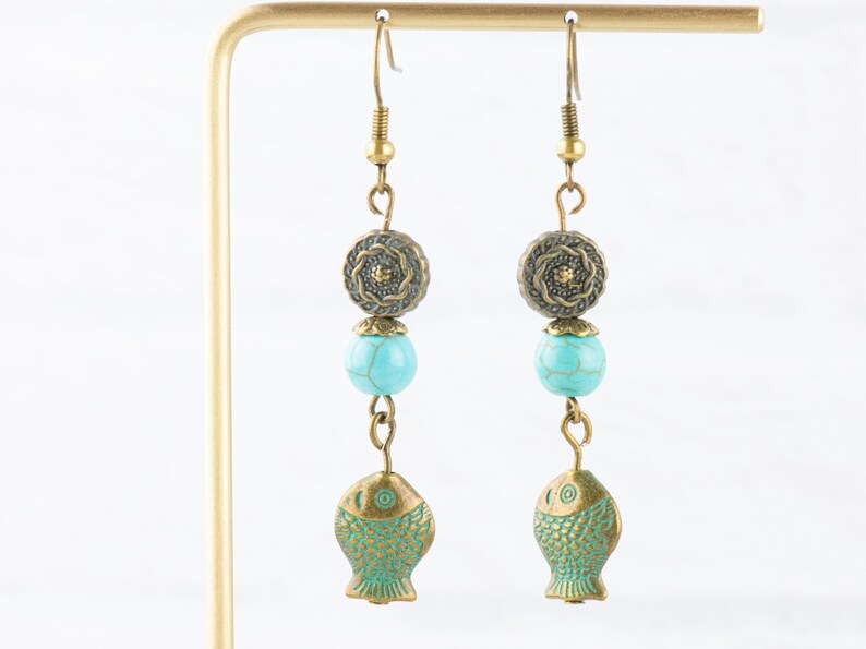 May include: A pair of turquoise and brass dangle earrings featuring a turquoise bead, a brass flower charm, and a brass fish charm.