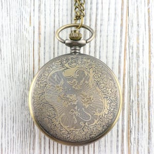 K&ouml;nnte beinhalten: Antike Taschenuhr mit detailliertem Blumendesign auf dem Deckel. Die Uhr hat ein bronzefarbenes Geh&auml;use und eine passende Kette. Der Hintergrund ist eine wei&szlig;e Holzoberfl&auml;che.