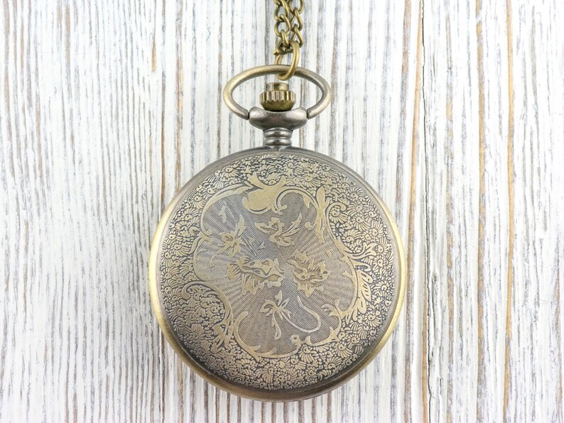 K&ouml;nnte beinhalten: Antike Taschenuhr mit detailliertem Blumendesign auf dem Deckel. Die Uhr hat ein bronzefarbenes Geh&auml;use und eine passende Kette. Der Hintergrund ist eine wei&szlig;e Holzoberfl&auml;che.