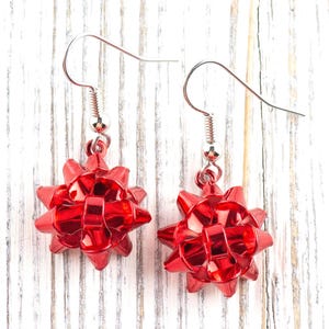 May include: A pair of red bow-shaped earrings. Each earring features a shiny, faceted bow design suspended from a silver-colored fish hook. The earrings are displayed against a white and gray wooden background.