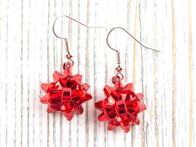 May include: A pair of red bow-shaped earrings. Each earring features a shiny, faceted bow design suspended from a silver-colored fish hook. The earrings are displayed against a white and gray wooden background.
