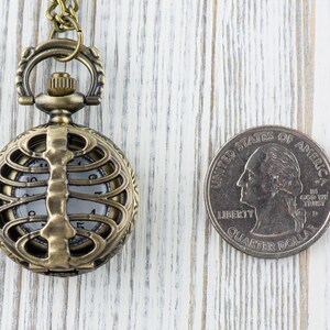 May include: A gold-toned pocket watch with a skeletonized design, featuring a ribcage pattern. The watch is hanging from a chain and is next to a US quarter.