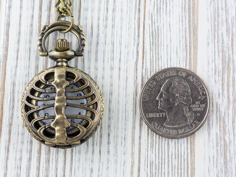 May include: A gold-toned pocket watch with a skeletonized design, featuring a ribcage pattern. The watch is hanging from a chain and is next to a US quarter.