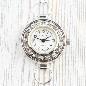 Könnte beinhalten: Eine silberfarbene Geneva-Quarzuhr mit weißem Zifferblatt und schwarzen Zeigern. Die Uhr hat eine dekorative Lünette mit kleinen, runden Verzierungen. Die Uhr hat zwei kreisförmige Schlaufen zur Befestigung.