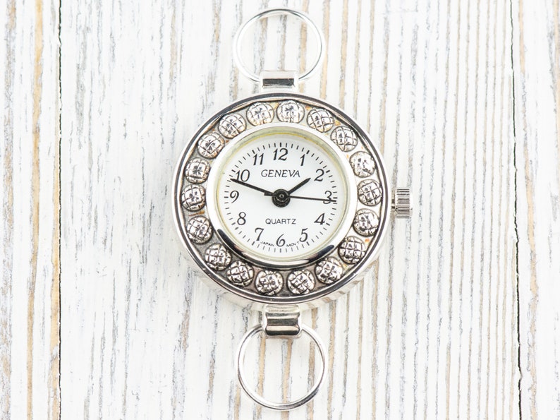 Pu&ograve; includere: Un orologio al quarzo Geneva argentato con quadrante bianco e lancette nere. L'orologio ha una lunetta decorativa con piccoli abbellimenti rotondi. L'orologio ha due anelli circolari per il fissaggio.