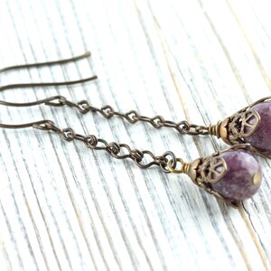 May include: A pair of handcrafted earrings with purple gemstone beads. The earrings have a bronze-coloured chain and decorative caps. The ear wires are bronze-coloured. The earrings are displayed on a white wooden surface.
