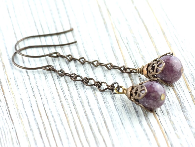 May include: A pair of handcrafted earrings with purple gemstone beads. The earrings have a bronze-coloured chain and decorative caps. The ear wires are bronze-coloured. The earrings are displayed on a white wooden surface.