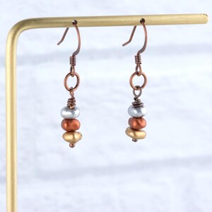 May include: A pair of handcrafted earrings with a copper-colored hook and wire. Each earring features a stack of three beads: silver, copper, and gold. The earrings are displayed on a gold-colored stand.