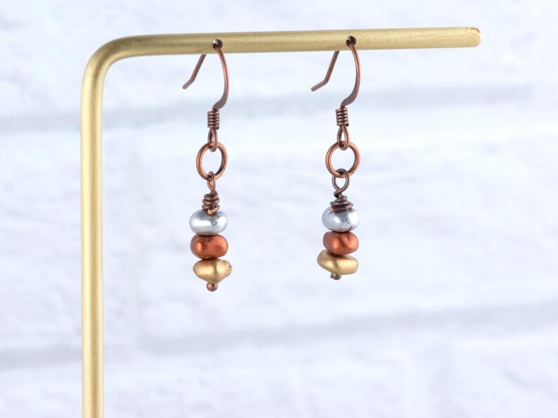 May include: A pair of handcrafted earrings with a copper-colored hook and wire. Each earring features a stack of three beads: silver, copper, and gold. The earrings are displayed on a gold-colored stand.