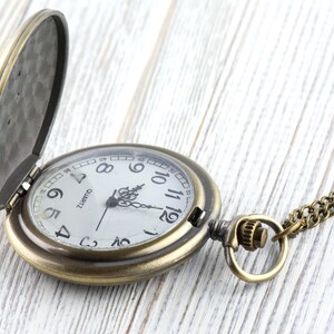 K&ouml;nnte beinhalten: Eine Taschenuhr im antiken Stil mit offenem Deckel, die ein wei&szlig;es Zifferblatt mit schwarzen Ziffern und Zeigern zeigt. Die Uhr hat eine Bronzefinish und eine passende Kette. Das Wort "QUARTZ" ist auf dem Zifferblatt aufgedruckt.