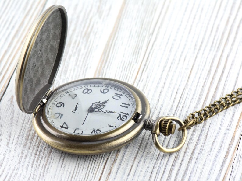 K&ouml;nnte beinhalten: Eine Taschenuhr im antiken Stil mit offenem Deckel, die ein wei&szlig;es Zifferblatt mit schwarzen Ziffern und Zeigern zeigt. Die Uhr hat eine Bronzefinish und eine passende Kette. Das Wort "QUARTZ" ist auf dem Zifferblatt aufgedruckt.