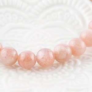 May include: A pink sunstone bracelet with round beads. The bracelet is on a white background.