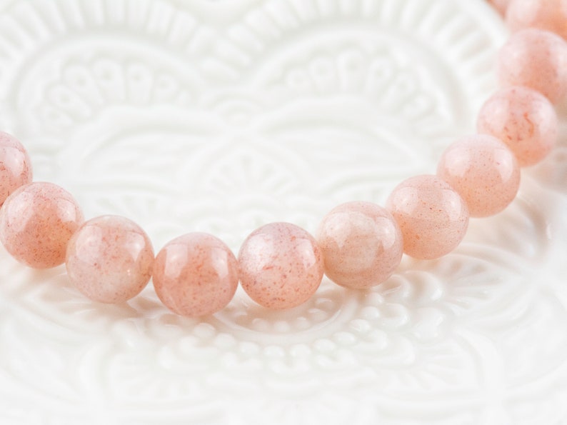 May include: A pink sunstone bracelet with round beads. The bracelet is on a white background.