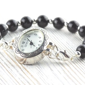 May include: A silver watch bracelet featuring a white watch face and a black beaded band. The watch face is marked with the word "GENEVA". The bracelet is displayed on a white wooden surface.