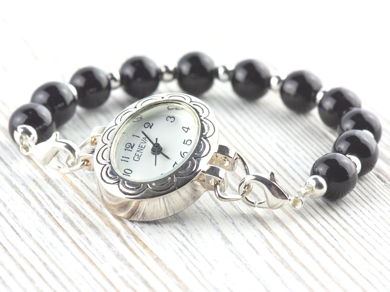May include: A silver watch bracelet featuring a white watch face and a black beaded band. The watch face is marked with the word "GENEVA". The bracelet is displayed on a white wooden surface.