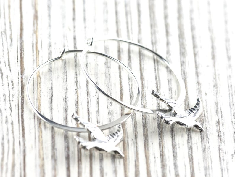 Peut inclure: Boucles d'oreilles cr&eacute;oles argent&eacute;es avec un charm en forme d'oiseau. Les boucles d'oreilles pr&eacute;sentent un design circulaire simple avec un petit pendentif oiseau d&eacute;taill&eacute;. Les oiseaux sont en vol, ajoutant une touche d'&eacute;l&eacute;gance inspir&eacute;e de la nature.
