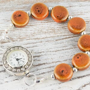 May include: A silver watch with a white face and a chain of orange ceramic beads. The watch face has a silver rim with small rhinestones around the edge. The watch face says "Geneve" and has black hands.