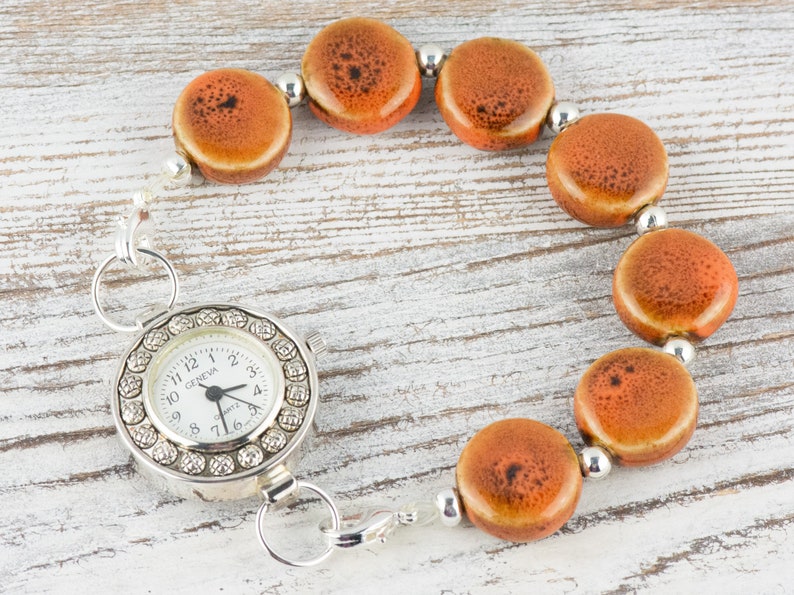 May include: A silver watch with a white face and a chain of orange ceramic beads. The watch face has a silver rim with small rhinestones around the edge. The watch face says "Geneve" and has black hands.
