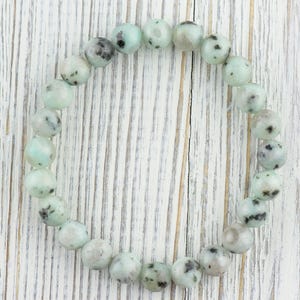 Puede incluir: Un brazalete hecho de cuentas redondas de color verde claro y negro moteado. Las cuentas son lisas y tienen un acabado pulido.