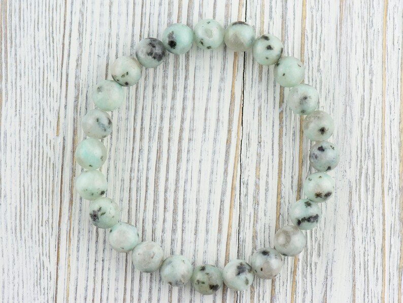 Puede incluir: Un brazalete hecho de cuentas redondas de color verde claro y negro moteado. Las cuentas son lisas y tienen un acabado pulido.