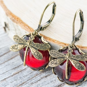 Op de afbeelding: Een paar rode glazen oorbellen met een libellenontwerp. De oorbellen hebben een gouden afwerking en zijn gemaakt met een haaksluiting.