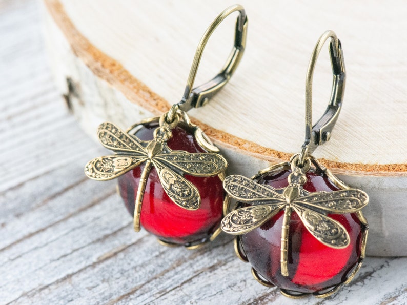 Pode incluir: Um par de brincos de vidro vermelho com um design de lib&eacute;lula. Os brincos t&ecirc;m um acabamento dourado e s&atilde;o feitos com fecho de gancho.
