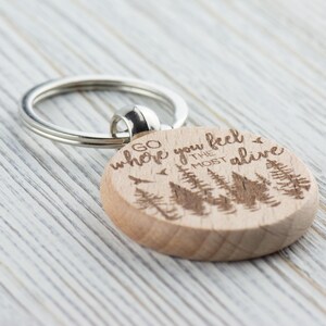 May include: Wooden keychain with a silver key ring. The keychain is engraved with the text "Go where you feel the most alive" and a silhouette of trees.