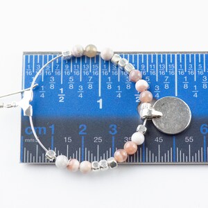 Pu&ograve; includere: Un orecchino a cerchio d'argento con un ciondolo e una fila di perline rosa e bianche. L'orecchino &egrave; misurato con un righello, mostrando che ha un diametro di 3 cm.